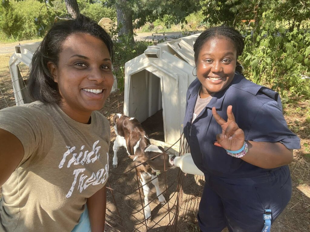 Two women pose for pictures while one is feeding a bottle to a brown and white calf