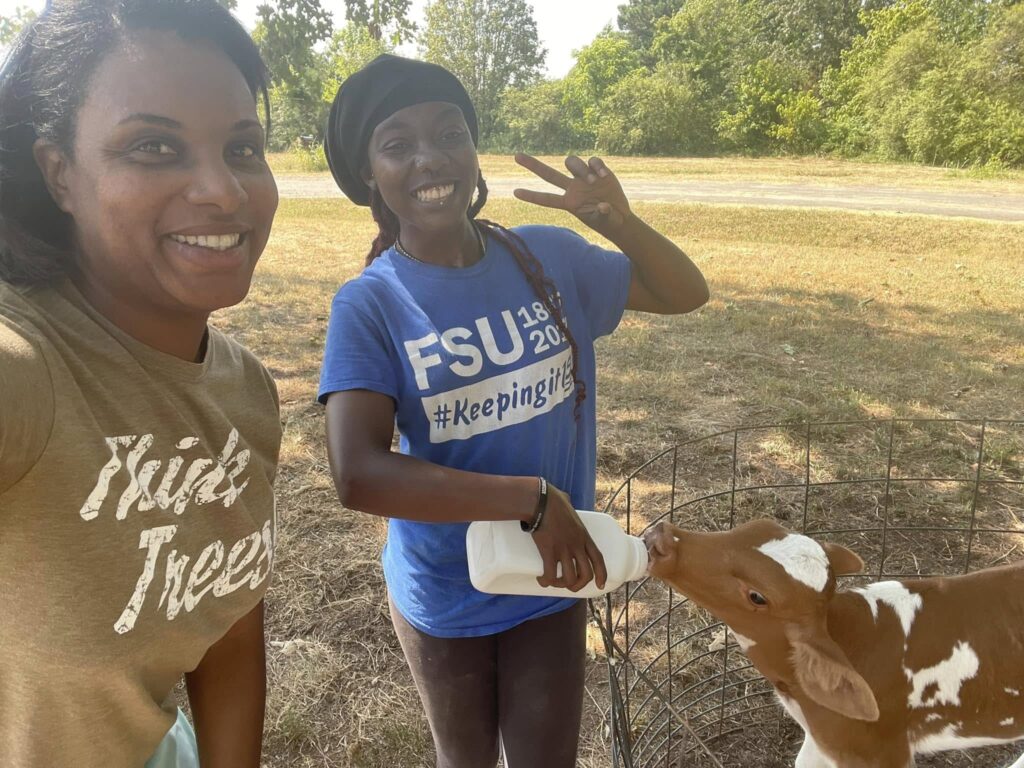 Two women pose while one is feeding a brown and white calf a bottle